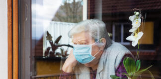 Elderly woman wearing mask, looking out window beside orchid.