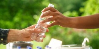 Hands passing a water bottle outdoors, lush green background.