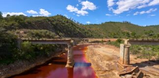 Weathered bridge over a red river with green hills.