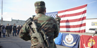 Soldier stands with rifle in front of American flag.