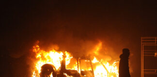 Burning vehicle at night, silhouetted person walking past.