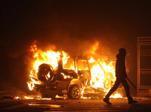 Burning vehicle at night, silhouetted person walking past.
