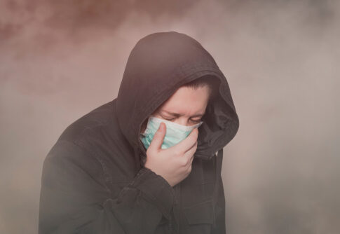 Person with hood and mask coughing in foggy setting.