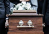 Pallbearers carrying a wooden coffin with white flowers.