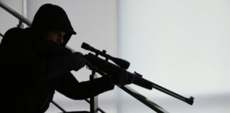 A silhouetted figure in tactical gear aiming a sniper rifle from a staircase