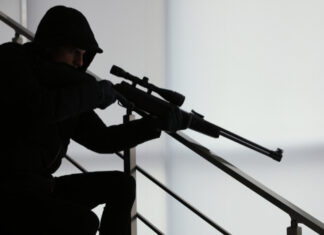 A silhouetted figure in tactical gear aiming a sniper rifle from a staircase