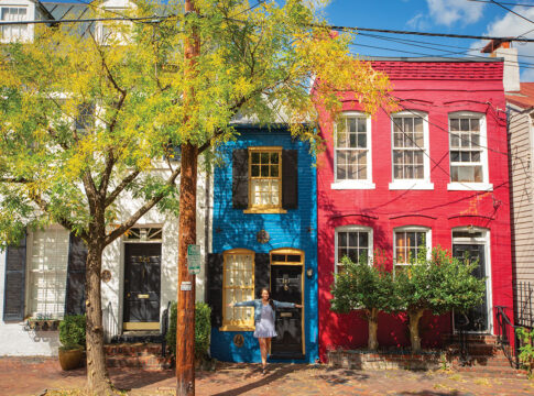 A narrow, colorful house flanked by a blue and a red house, with a person standing in front, surrounded by autumn trees.