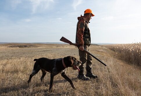 A man in hunting gear walking with a dog in a field during sunset