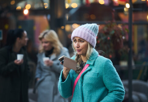 A woman in a blue coat and pink beanie speaking into a smartphone in an outdoor setting