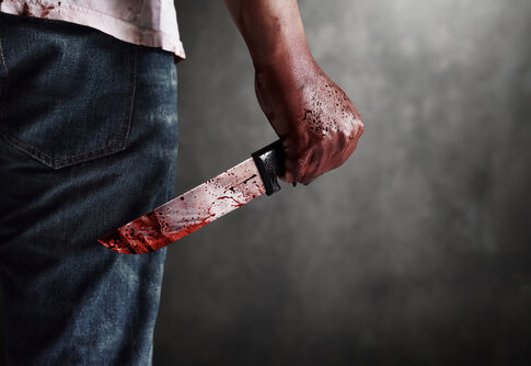 A man holding a bloody knife behind his back in a dark setting
