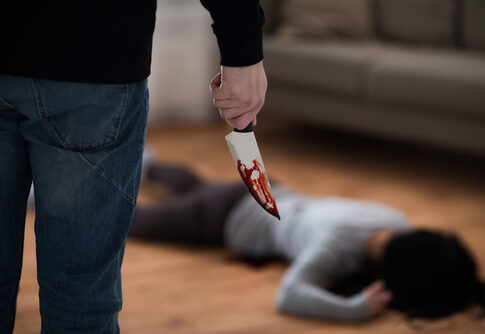 A person holding a bloody knife stands over a victim lying on the floor